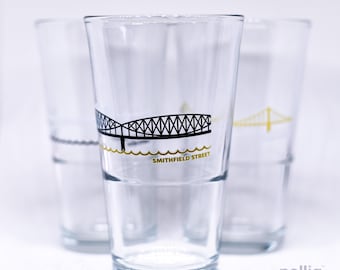 May include: Three clear glass tumblers featuring a black graphic of the Smithfield Street bridge above a gold wavy line. The text SMITHFIELD STREET is printed in gold below the bridge graphic on the front-most glass, with a white background.