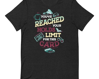 May include: Dark grey t-shirt featuring the text "You've Reached Your Holds Limit For This Card." The design includes a coffee cup, books, a library card, and glasses. The shirt is a short-sleeved crew neck.