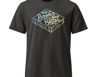 May include: A charcoal grey short-sleeve t-shirt featuring a graphic of a book with the text "THE BOOK WAS BETTER" written in a stylised, gradient font that transitions from light blue to yellow across the cover of the illustrated book.
