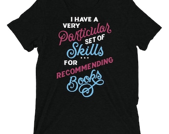 May include: A black short-sleeved t-shirt featuring the white, pink, and light blue text: I HAVE A VERY Particular SET OF Skills FOR RECOMMENDING Books. The design uses a mix of bold sans-serif and decorative script fonts centred on the chest.