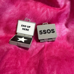 May include: Two small gray plastic briefcase-shaped items. One is open, revealing a white interior with the black text END UP HERE and a black base with a white star. The other is closed, displaying the black text 5SOS with a star in the middle.
