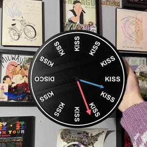May include: A black, round decorative clock with the word "KISS" repeated around the edge and "DISCO" at the top. The clock has blue and red hands. The clock is held in front of a wall with framed art and posters.