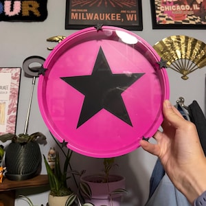 Puede incluir: Una bandeja circular con un borde rosa brillante y un centro transparente que presenta un gráfico de una estrella negra grande y sólida. El borde exterior del marco rosa está decorado con cuatro pequeños detalles de estrellas negras en 3D.