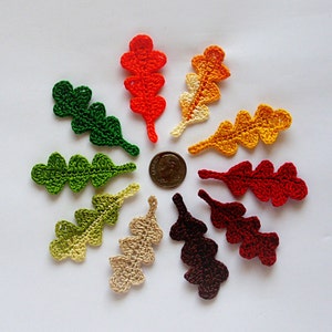 Puede incluir: Una colección de apliques de hojas de roble de ganchillo en varios colores otoñales, incluyendo verde, naranja, amarillo y marrón. Las hojas están dispuestas en un patrón circular, con una moneda en el centro para referencia de tamaño. Estas hojas hechas a mano son ideales para manualidades.