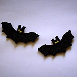 May include: Two black, handmade crochet bats with yellow eyes. The bats have a wing span design and are perfect for Halloween decorations or craft projects. The bats are displayed on a white background.