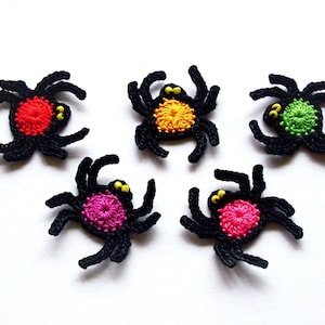 Pode incluir: Cinco aranhas pretas de crochê com centros de cores diferentes. As aranhas têm olhos amarelos e estão dispostas em círculo sobre um fundo branco.