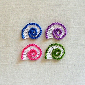 Puede incluir: Cuatro conchas de mar de ganchillo en diferentes colores. Las conchas son blancas con una espiral de azul, rosa, verde y morado.