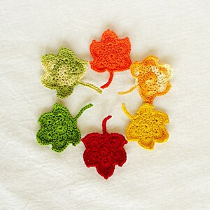 May include: Six crocheted maple leaf shapes in various shades of green, orange, and red. The leaves are arranged in a circle on a white background.