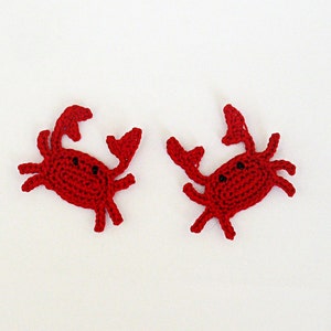 May include: Two red crocheted crabs with black eyes. The crabs are facing each other and are on a white background.