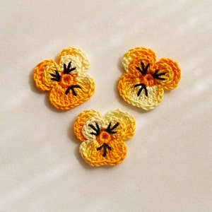 May include: Three crocheted yellow and white flowers with black centers. The flowers are in the shape of pansies.