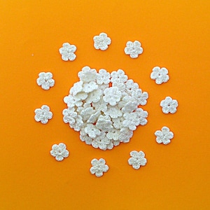 May include: A collection of small, off-white crochet flowers arranged on a bright orange background. The flowers are clustered in the center and scattered around the edges, creating a decorative pattern. The flowers are approximately 2.5 cm in diameter.