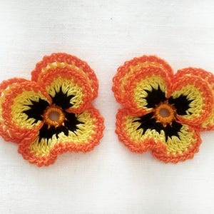 May include: Two handmade crochet pansy flowers. The flowers feature a vibrant color palette of orange, yellow, and black. The petals are orange, transitioning to yellow, with a black center. These decorative flowers are perfect for craft projects.