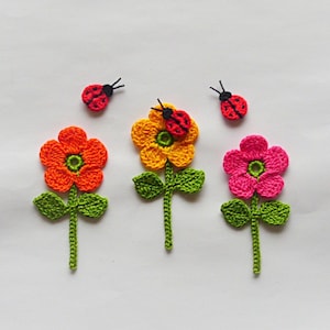 Puede incluir: Tres flores de ganchillo con mariquitas en la parte superior. Las flores son naranja, amarilla y rosa. Las mariquitas son rojas con manchas negras.