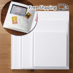 Puede incluir: Tres álbumes de fotos blancos cuadrados en blanco apilados uno encima del otro. El álbum superior es ligeramente más pequeño que los dos álbumes inferiores. Los álbumes están sobre una superficie de madera.