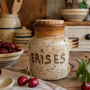 May include: A speckled beige ceramic jar with a cork lid and a side handle, featuring the embossed text CERISES. The jar is surrounded by fresh red cherries with green stems, a small ceramic spoon, and a beige cloth on a wooden surface.