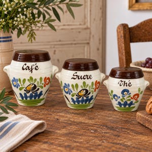 May include: A set of three cream-colored ceramic canisters with brown lids and side handles. Each canister features hand-painted floral motifs in blue, green, and orange, with the words Café, Sucre, and Thé written in black script on the front.