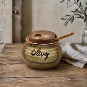 May include: A round ceramic jar with a tan and brown gradient glaze, featuring the word "Olives" written in dark script. The jar has a matching brown lid with a small circular cutout and a wooden spoon resting inside the opening.