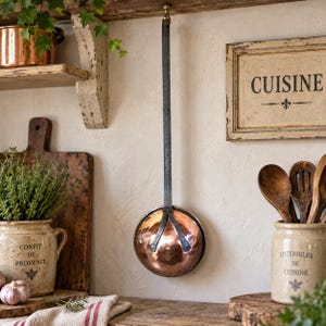 May include: A long-handled kitchen ladle featuring a hammered copper bowl and a dark, flat iron handle. The ladle is suspended from a hook, showcasing a metallic finish with a rounded basin and a sturdy, vertical metal support structure.