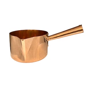 May include: A copper-colored saucepan with a long, angled handle. The pan has a rounded body with a pouring spout. The handle is attached to the side of the pan. The interior of the pan is a darker copper color.