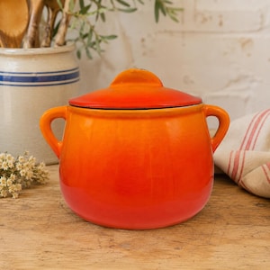 May include: A round, orange and red gradient ceramic pot with two side handles and a matching lid. The pot features a smooth, glossy finish that transitions from a deep red at the base to a bright orange at the top, with a rounded lid on top.