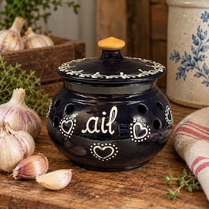 May include: A dark blue ceramic garlic keeper with a lid, featuring white hand-painted heart motifs and the word "ail" in cursive script. The round container has multiple circular ventilation holes around the upper body and a small tan knob.