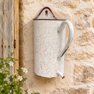 May include: A wall-mounted white enamel pitcher featuring a red marbled pattern, a curved handle, and a small spout at the base. The top edge is trimmed in dark red, and the back plate includes a hole for hanging on a wall surface.