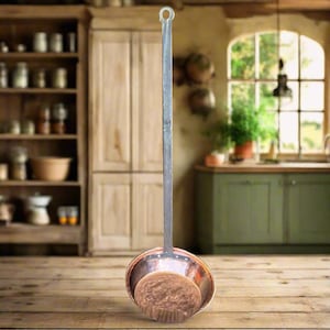 May include: A vintage copper and metal ladle with a long handle and a round, bowl-shaped head. The ladle has a rustic, aged appearance, suggesting it is an antique kitchen tool. The handle has a loop at the end for hanging.