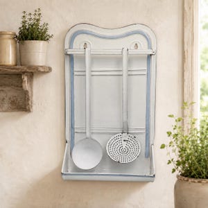 May include: A white enamelled metal wall-mounted kitchen rack with a blue trim border. The rack features a horizontal bar for hanging utensils, a bottom shelf, and two hanging kitchen tools: a solid round ladle and a perforated skimmer spoon.