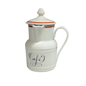 May include: White ceramic coffee pot with a lid and handle. The pot has a black "Cafe" script design on the front, with a red and black stripe around the top. The pot is designed for serving coffee.