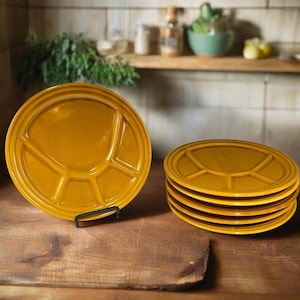 May include: A stack of six golden-yellow divided plates, with a single plate displayed on a black stand. The plates have a circular design with three compartments. The plates are on a wooden surface.