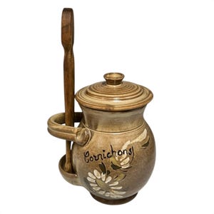 May include: A ceramic jar with a lid and a wooden spoon. The jar is tan with a floral design and the word "Cornichons" written on it. The spoon rests in a holder attached to the side of the jar.