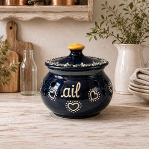 May include: A dark blue ceramic garlic keeper with a rounded body, a matching lid with a yellow knob, and ventilation holes. The word "ail" is written in cream-coloured script on the front, surrounded by cream-coloured heart outlines and dots.