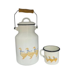 May include: A white enamel milk jug and mug set. The jug has a wooden handle and a black metal handle. Both pieces are decorated with a repeating pattern of yellow ducks wearing blue bows.