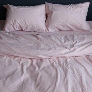 May include: A light pink bed set, including two pillowcases and a fitted sheet. The bedding is made of a soft, wrinkled fabric, and the color is a pale, muted pink. The background is a dark blue-gray.