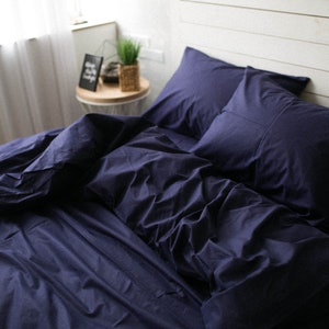 May include: A navy blue bed set, including a duvet cover and two pillowcases, arranged on a bed. The bedding is made of a soft fabric and has a slightly wrinkled appearance. A small side table with a framed picture and a plant is visible in the background.