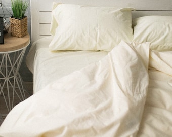 May include: Cream-colored bed linens, including a pillow and a duvet cover, are arranged on a bed. The bedding is made of a soft, possibly cotton, material. The scene is lit with natural light, creating a cozy and inviting atmosphere. A small side table with a plant is visible.