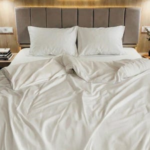 May include: A bed dressed with white bedding, including a duvet cover and two pillows. The bed is made up. The headboard is a neutral colour, and the room has wooden accents.