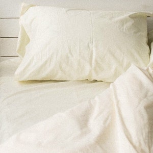 May include: Cream-colored bed linens, including a pillow and a folded duvet, arranged on a bed. The bedding is made of a soft, possibly cotton, material. The neutral color palette suggests a focus on comfort and simplicity in the bedroom.