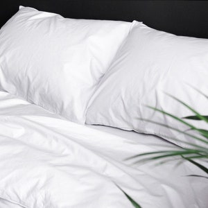 Puede incluir: Primer plano de una cama con ropa de cama blanca, que incluye dos almohadas y una s&aacute;bana bajera. La ropa de cama est&aacute; cuidadosamente colocada, con ligeras arrugas que a&ntilde;aden textura. Algunas hojas verdes de plantas est&aacute;n en primer plano.