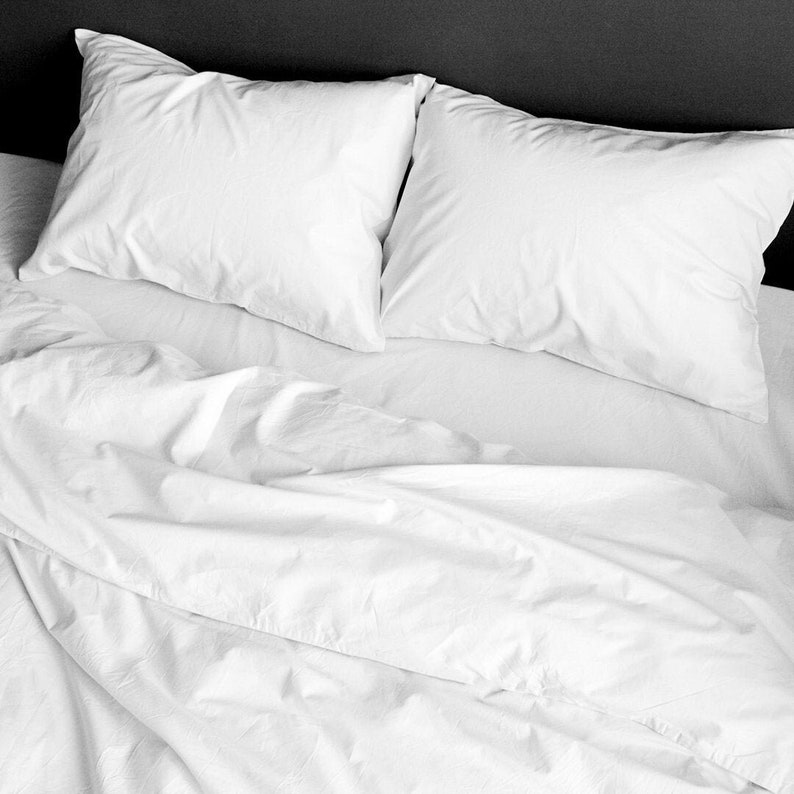Puede incluir: Primer plano de una cama con ropa de cama blanca. Dos almohadas blancas y esponjosas descansan sobre una s&aacute;bana blanca. La s&aacute;bana y la manta est&aacute;n ligeramente arrugadas, creando un aspecto suave y acogedor. El fondo es gris oscuro.