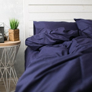 May include: A navy blue bed set with a duvet cover and pillowcases. The bedding is made of a soft fabric and is neatly arranged on a bed with a white headboard. A small side table with a plant and a decorative frame is in the background.