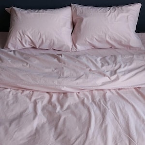 May include: A light pink bed set comprising a duvet cover and two pillowcases. The bedding, made from a soft, slightly creased fabric, gives a relaxed appearance. The set is arranged neatly, suggesting a focus on bedroom decor and comfort.