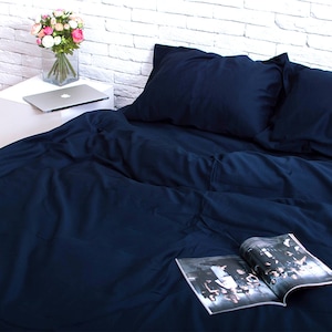 May include: A navy blue bed set, including a duvet cover and pillowcases, is arranged on a bed. An open magazine lies on the bed. A vase of flowers and a laptop sit on a white bedside table.