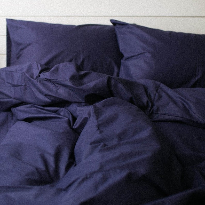 May include: A navy blue bed set, including a duvet cover and two pillowcases, is arranged on a bed. The bedding is made of a smooth fabric and has a slightly rumpled appearance. The background is a white wooden wall.