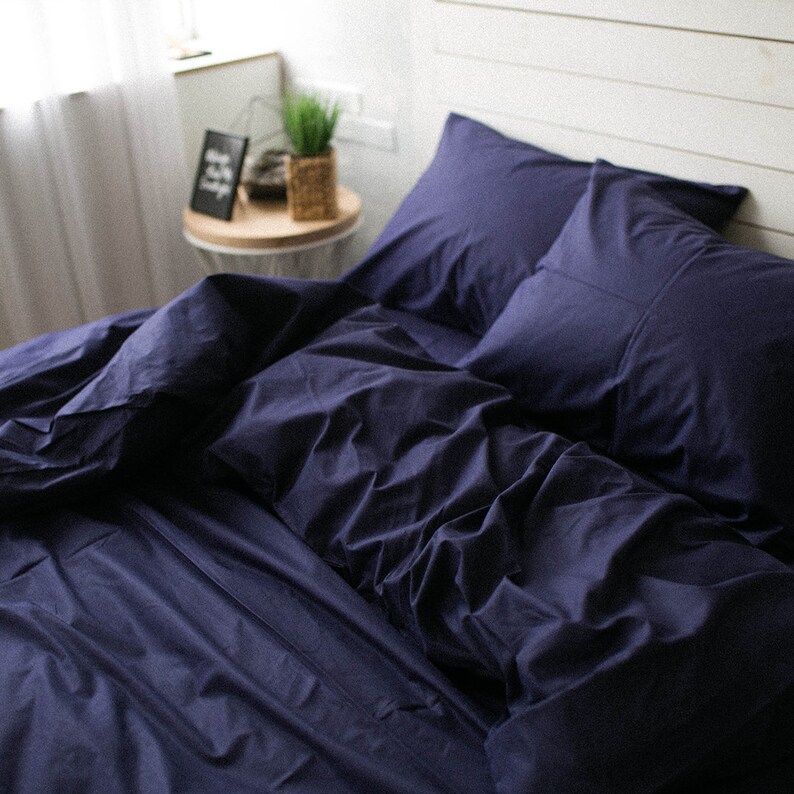 May include: A navy blue bed set, including a duvet cover and two pillowcases, arranged on a bed. The bedding is made of a soft fabric and has a slightly wrinkled appearance. A small side table with a framed picture and a plant is visible in the background.