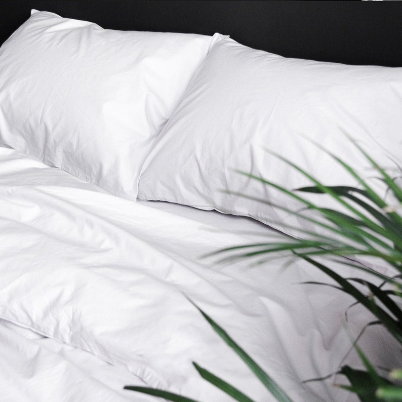 Puede incluir: Primer plano de una cama con ropa de cama blanca, que incluye dos almohadas y una s&aacute;bana bajera. La ropa de cama est&aacute; cuidadosamente colocada, con ligeras arrugas que a&ntilde;aden textura. Algunas hojas verdes de plantas est&aacute;n en primer plano.