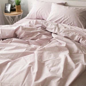 May include: A bed dressed with light pink bed linen, including a duvet cover and two pillowcases. The sheets are creased, suggesting recent use. The bed is against a white headboard. A small bedside table with a plant and picture frame is visible.