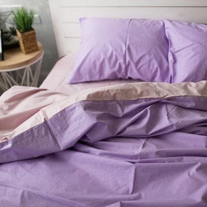 May include: A bed made with lavender-coloured bedding, including a pillow and a folded sheet. The sheet has a light pink border. The bed is made up and ready for use. A small potted plant is visible in the background.