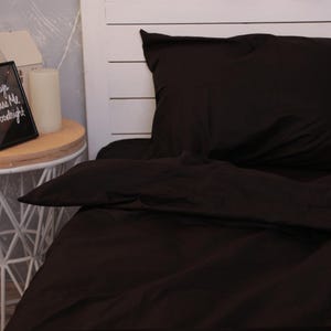 May include: A dark brown bed set, including a pillow and a sheet, is displayed on a bed. The bedding is made of a smooth fabric. A white headboard is visible in the background. A small side table with a candle and a framed picture is on the left.