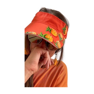 May include: A red visor with a white trim and a pineapple print. The visor is worn by a person with brown hair and sunglasses.